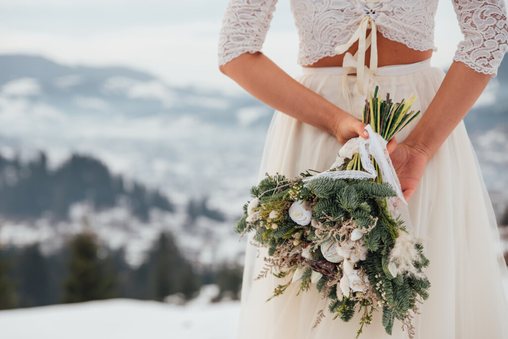Bride in white wedding dress holding colorful flowers bouquet in hands and posing outdoors. Winter wedding and season floral concept. Mountains on background.