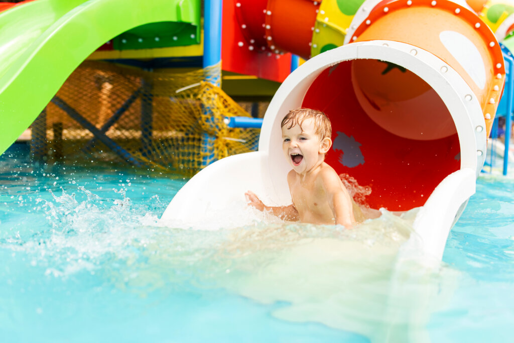 Happy laughing little boy playing on water slide in outdoor swimming pool on a hot summer day. Child sliding on aqua playground in tropical resort