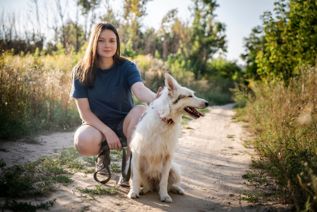 The girl looks into the camera and a white dog is sitting next to her on a dirt road against the background of a forest in the daytime.