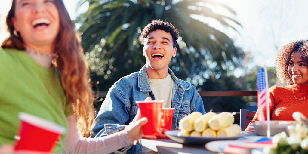 Friends, celebration and independence day in USA with backyard food, drinks and international holiday. Social group of people with American Flag, barbecue and lunch at patio table.