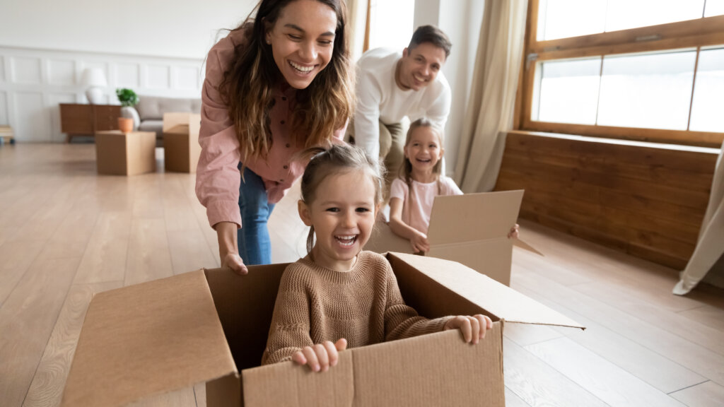 Happy little girls sisters sitting in big carton boxes while joyful parents pushing it on floor. Laughing family couple having fun with small children in new house, relocation moving day concept.